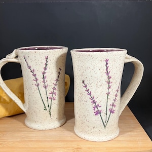 May include: Two handmade ceramic mugs with a speckled, off-white finish. Each mug features a hand-painted lavender sprig design in purple and green. The interior and rim of the mugs are a deep purple. The mugs are on a wooden surface.