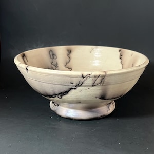 May include: A round, off-white ceramic bowl featuring a smooth, glossy finish and distinct, dark, branching lightning-like patterns that wrap around the exterior and interior surfaces. The bowl sits on a raised, circular pedestal base.