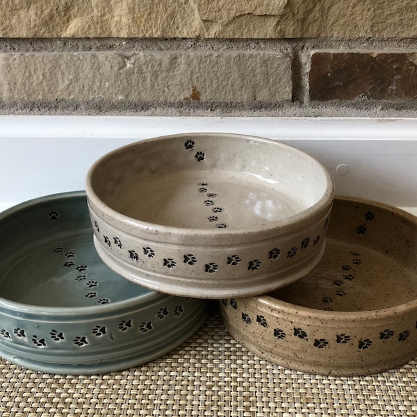 May include: Three ceramic pet bowls with paw print designs. The bowls are in shades of beige, light brown, and blue-grey.