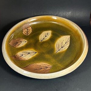 May include: A round ceramic serving dish with a green and yellow interior and a cream-colored rim. The dish is decorated with brown and tan leaf designs. The dish is likely used for serving food.