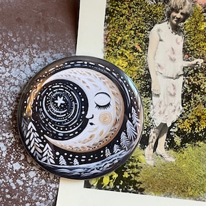 May include: A black and gold moon and stars design on a round button. The button features a stylized moon with a closed eye and a star in the center. The moon is surrounded by a ring of stars and a border of trees.