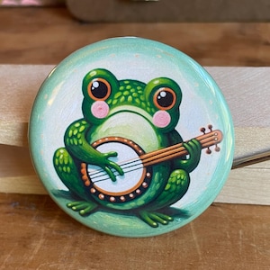 May include: A round button with a cartoon frog playing a banjo. The frog is green with large eyes and a pink blush. The button has a light green border and a white background.