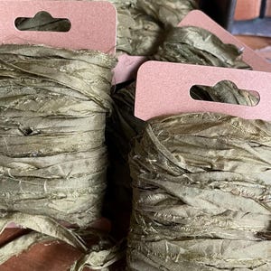 May include: Spools of olive green silk ribbon with frayed edges, each attached to a brown tag. The ribbon is tightly wound, with some spools partially unwound. A metal container holds additional spools in the background.