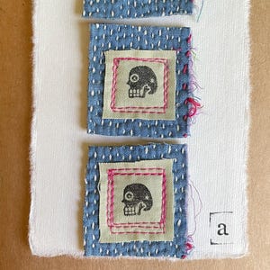 May include: Three square fabric patches featuring black skull prints on light green centers, framed by blue fabric with white sashiko-style stitching and pink thread accents. The patches are mounted on a white card with the letter a and text.