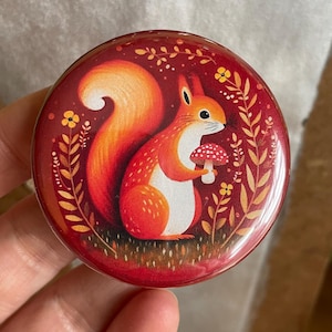 May include: A round button with a red background featuring a cartoon squirrel holding a mushroom. The squirrel has orange fur and a white belly. The button has a glossy finish and is surrounded by floral elements.