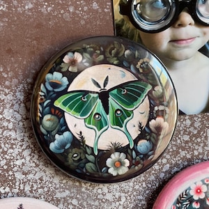 May include: A round, black, magnetic pin with a green luna moth surrounded by white and blue flowers on a dark background.