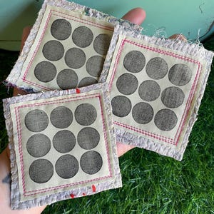 May include: Three square fabric coasters featuring a cream-colored center panel with a grid of nine dark gray circles. Each coaster is bordered by a bright pink stitched frame and finished with a textured, frayed, light-colored fabric edge.