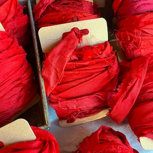 May include: Close-up of several spools of bright red fabric ribbon. Each spool is neatly wound and attached to a small, rectangular cardboard tag. The ribbon has a slightly frayed texture, suggesting a handmade or vintage quality. The spools are arranged in a metal container.