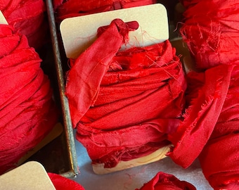 May include: Multiple spools of vibrant red fabric ribbon wrapped around rectangular cardboard cards. The ribbon features a raw, frayed edge texture and a deep crimson colour, with each spool neatly wound to display the layered fabric material.