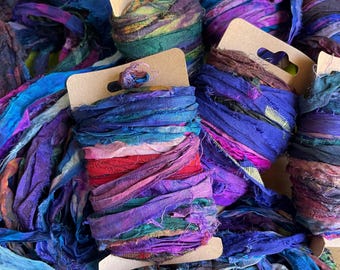 May include: A collection of colorful silk ribbon spools in shades of blue, purple, and red. The spools are displayed on cardboard tags. The ribbons have a textured, frayed appearance, suggesting a handmade or vintage quality. Ideal for crafting and textile projects.