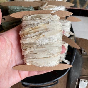 May include: Cream-colored ribbon wound on a wooden spool. The ribbon has a frayed, textured appearance. Several spools of ribbon are visible, some in shades of green and cream. The spools are arranged in a metal container.