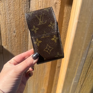 May include: Brown leather case with a repeating pattern of gold-colored interlocking letters and floral shapes. The case is rectangular and has a flap closure. The case is held in a hand.