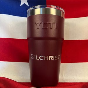 May include: A maroon insulated tumbler with the brand name "YETI" embossed on the front. The name "GILCHRIST" is printed below. The tumbler has a silver rim and is set against a backdrop of a red, white, and blue flag.