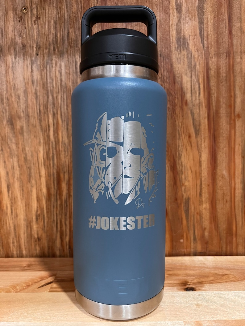 May include: A blue Yeti water bottle with a stainless steel lid and a black handle. The bottle features a laser-etched design of a horror movie character with the text "#JOKESTER" underneath.