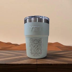 May include: A light sage green YETI tumbler with a clear plastic lid and a stainless steel rim. The front features an engraved crest with a sword and dragonfly, along with the text "James Kelley Bell" and the date "May 30, 2021" below the design.