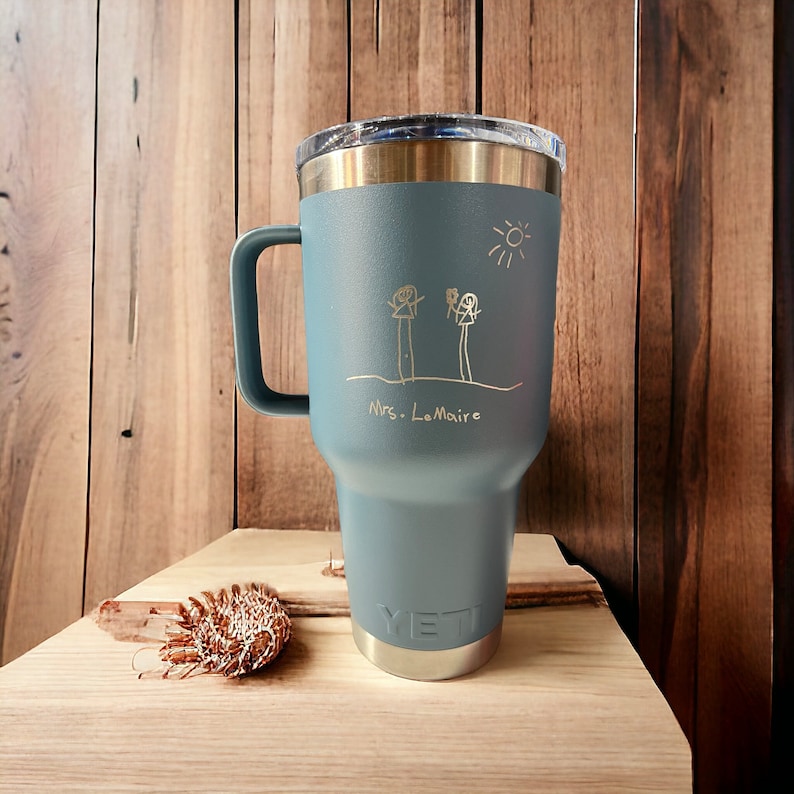 May include: A blue Yeti tumbler with a gold inscription that reads "Mrs. LeMaire" and a child's drawing of two stick figures and a sun.