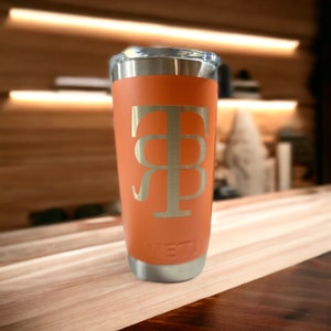 May include: A peach-coloured stainless steel tumbler with a gold monogram "T&B" on the side. The tumbler is sitting on a wooden surface.