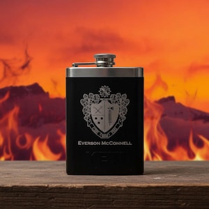 May include: Black stainless steel flask with a silver cap. The flask features an engraved crest design with the name "Everson McConnell" and the brand "YETI" at the bottom. The background shows a fiery orange and red gradient.