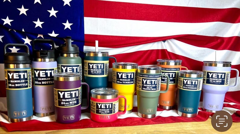 May include: A collection of YETI brand insulated drinkware in various sizes and colours, including tumblers, mugs, and bottles. The drinkware is arranged on a red, white and blue American flag background.