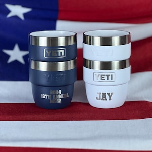May include: Two insulated tumblers, one navy blue and one white, each with a silver rim and the "YETI" logo. The navy tumbler has "2024 18TH ANNUAL MSW" printed on it, and the white tumbler has "JAY" printed on it. The background is an American flag.