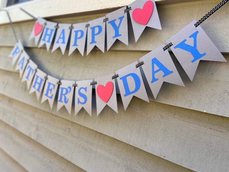 May include: A brown paper banner with the words "Happy Father's Day" in blue letters and red hearts. The banner is strung with black and white ribbon.