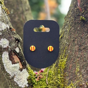 May include: A pair of small, yellow and red softball earrings on a black card with a hole for hanging.