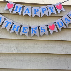 May include: A gray banner with the words "Happy Father's Day" in blue letters and red hearts. The banner is strung up on a white wooden wall.