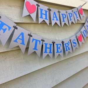 May include: A gray and blue banner with the text "Happy Father's Day" and red hearts. The banner is strung on a black and white string.