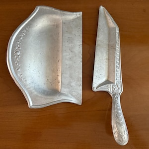 May include: A silver-colored cake server and matching serving plate. The server has a decorative handle and a blade with a geometric design. The plate has a curved shape with floral detailing along the edge. The set is displayed on a wooden surface.