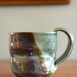 May include: Handmade ceramic mug with a unique, organic shape. The mug features a two-tone design with brown and green glaze, creating a rustic and earthy aesthetic. The handle is curved and integrated into the mug's body. A perfect addition to any kitchen.