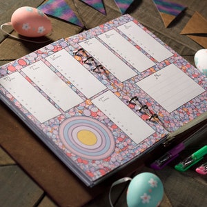 Könnte beinhalten: Ein brauner ledergebundener Planer mit einem farbenfrohen Muster auf dem Einband. Der Planer hat eine Ringbindung und ist auf eine Wochenübersicht geöffnet. Neben dem Planer liegen drei Stifte auf dem Tisch.
