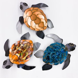 Puede incluir: Tres esculturas de tortugas de metal en diferentes colores. Las tortugas están en una variedad de poses, con la cabeza levantada y las patas extendidas. Las tortugas están pintadas en tonos de naranja, azul y marrón.