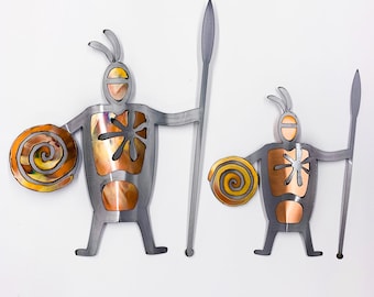 May include: Two metal wall art pieces depicting stylized figures. The figures are gray with copper and gold accents, holding spears and shields. The shields have a spiral design. The figures have a unique, abstract design, suitable for home decor.