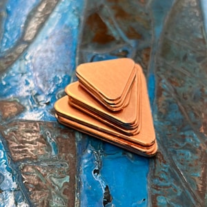 May include: A stack of four copper triangle shapes, each with a slightly different size, arranged in a pyramid shape on a blue and green textured surface.