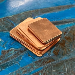May include: A stack of six square copper sheets in various sizes, with the largest sheet on top. The sheets are arranged in a staggered pattern, creating a layered effect. The copper has a warm, reddish-brown color and a slightly textured surface.