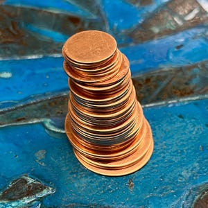 May include: A stack of round copper discs, each with a slightly different diameter, arranged in a cone shape on a blue and green textured surface.