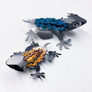 May include: Two metal lizards, one with a blue and green patterned back and the other with a gold and brown patterned back. The lizards are attached to each other and are made of metal.