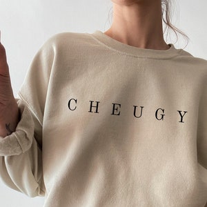 May include: A beige sweatshirt with the word "CHEUGY" printed in black on the front. The sweatshirt is being worn by a person wearing light blue jeans with distressed details.
