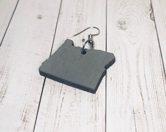 May include: A gray wooden earring shaped like the state of Oregon. The earring has a silver hook.