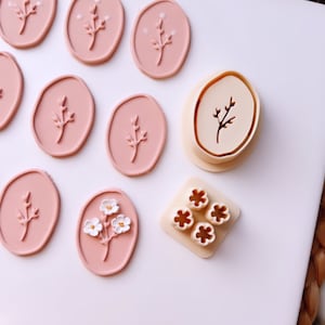 Puede incluir: Un conjunto de sellos de arcilla polimérica rosa con diseños florales. El conjunto incluye un sello con una sola rama de flores y un sello con cuatro flores individuales. Los sellos están sobre una superficie blanca.