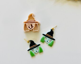 Op de afbeelding: Een Halloween-sieradenset met een heksenontwerp. De set bevat twee oorbellen en een koekjesvorm. De oorbellen hebben een heksengezicht met groen haar, een zwarte hoed en goudkleurige oorbelpalen. De koekjesvorm heeft de vorm van een heks.