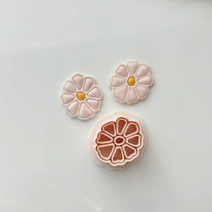 Puede incluir: Tres cortadores de galletas con forma de flores. Dos son rosas con un centro amarillo y uno es marrón con un centro blanco.