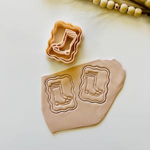 Peut inclure: Un ensemble de découpoirs à biscuits marron clair représentant des bottes de pluie. L'ensemble comprend un découpoir et deux empreintes sur une pâte. La pâte est marron clair. Le découpoir est de forme carrée ondulée.