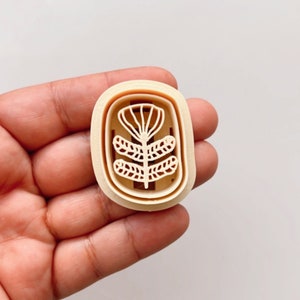 Puede incluir: Un cortador de galletas de plástico beige con forma de planta estilizada con tres hojas. El cortador tiene un borde elevado alrededor del exterior.