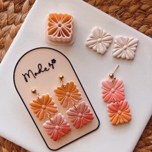 Puede incluir: Un par de pendientes hechos a mano de arcilla polimérica con un diseño de flores superpuestas en tonos naranja, rosa y amarillo. Los pendientes se muestran en una tarjeta blanca con el texto "Made by" en ella. La imagen también muestra dos cortadores de galletas con forma de flor.