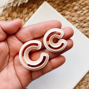Puede incluir: Dos cortadores de galletas de plástico en forma de la letra "C". Los cortadores son de color beige claro y se sostienen en una mano.