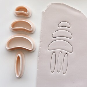 May include: A collection of five pale pink polymer clay cutters in different curved forms, alongside a piece of clay with the same design imprinted. These cutters are for creating abstract shapes for jewellery or other crafts.