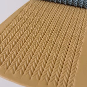 May include: A textured rolling pin with a leaf pattern is rolling across a tan surface, creating a repeating braided design. The rolling pin is a muted teal color. The tan surface is a rectangular shape.