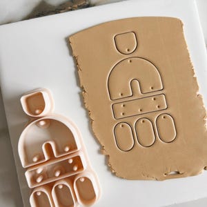 May include: A pink plastic cookie cutter shaped like a house with a roof, a door, and windows. The cutter is on a white surface next to a brown piece of clay with the same shape cut out.