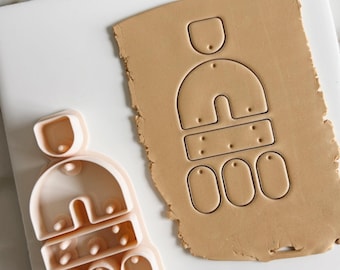 Op de afbeelding: Een roze plastic koekjesvormpje in de vorm van een huis met een dak, een deur en ramen. De vormpje ligt op een wit oppervlak naast een bruin stuk klei met dezelfde vorm uitgesneden.
